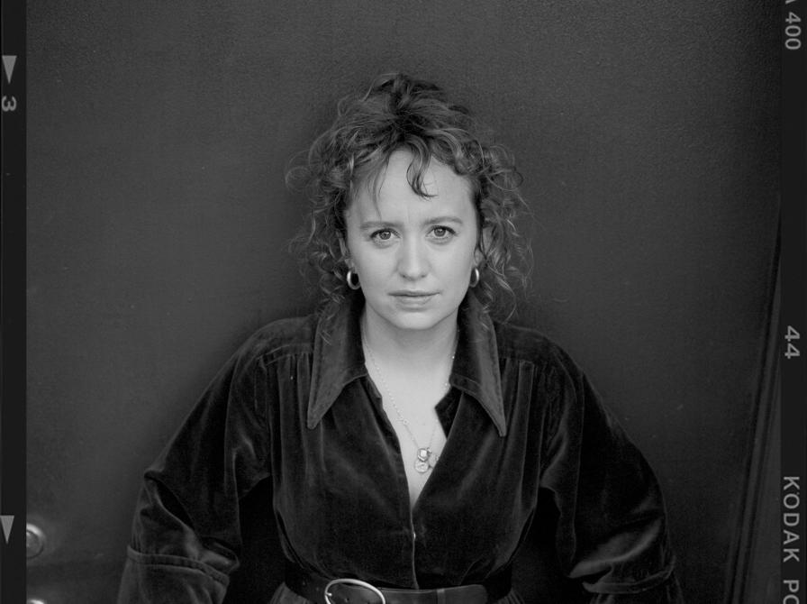 Sort-hvidt portræt af forfatteren Olga Ravn i mørk kjole med bælte, stående foran en mørk baggrund.